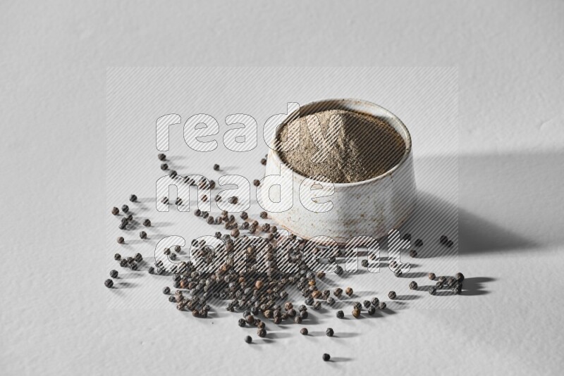 A white ceramic bowl full of black pepper powder and black pepper beads spread on white flooring