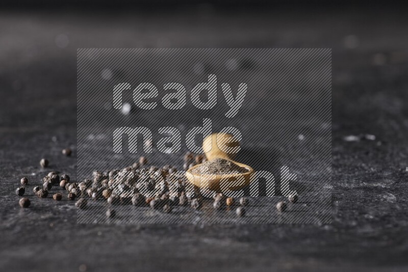A wooden spoon full of black pepper powder and black pepper beads spread on a textured black flooring