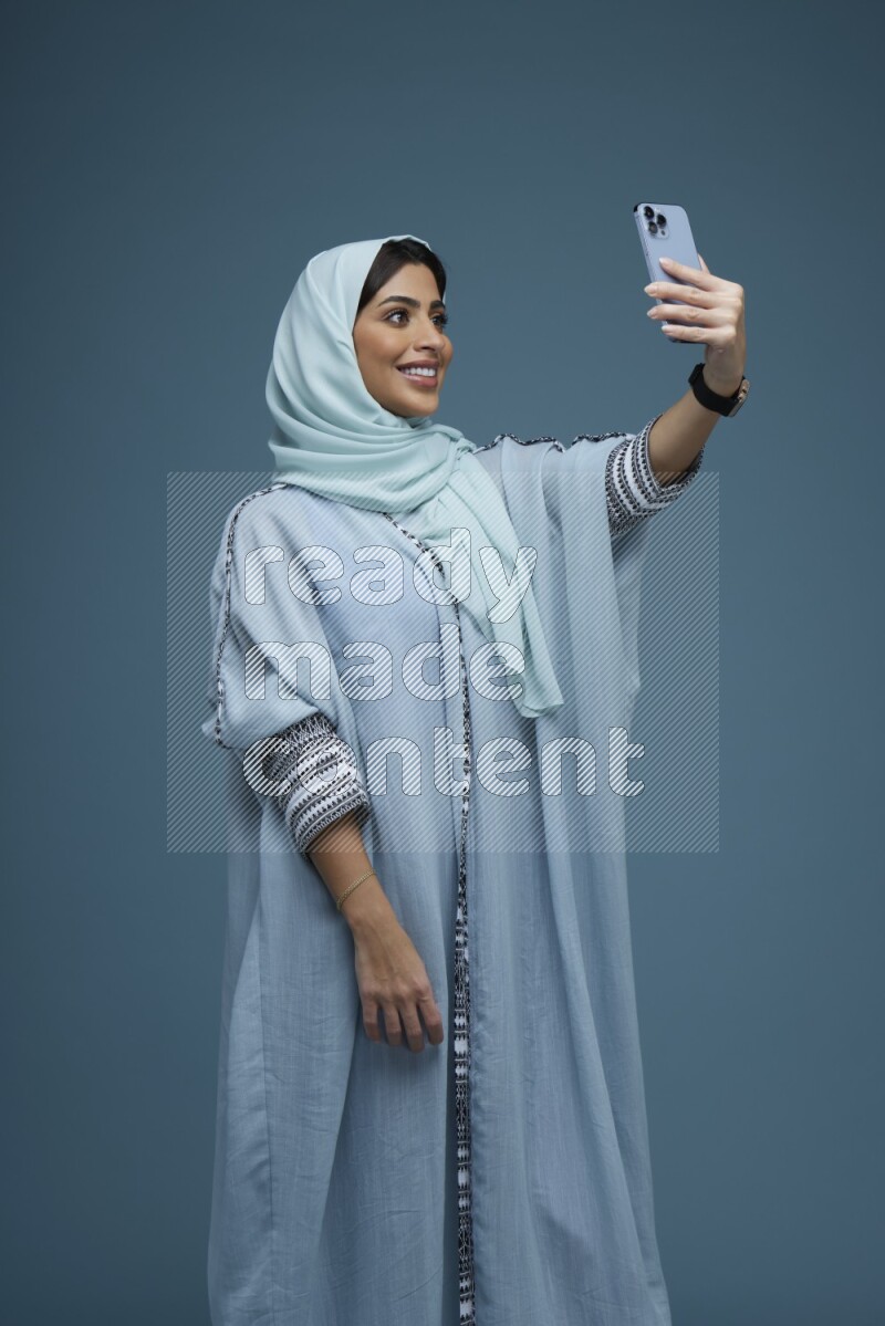A Saudi woman Taking a Selfie on a blue background wearing a blue Abaya with hijab