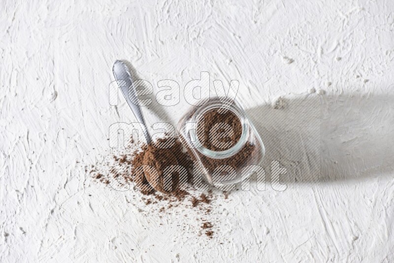 A glass spice jar and a metal spoon full of cloves powder on textured white flooring