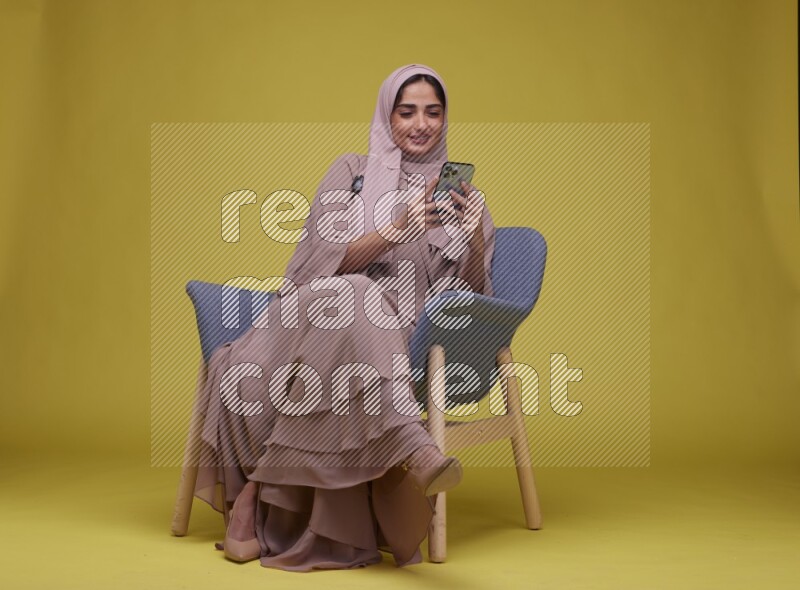A Female Texting Sitting  on a Yellow Background wearing Brown Abaya with Hijab
