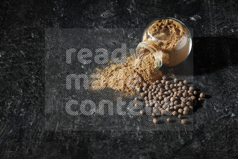A flipped glass spice jar full of allspice powder and powder spilled out of it with whole balls on a textured black flooring