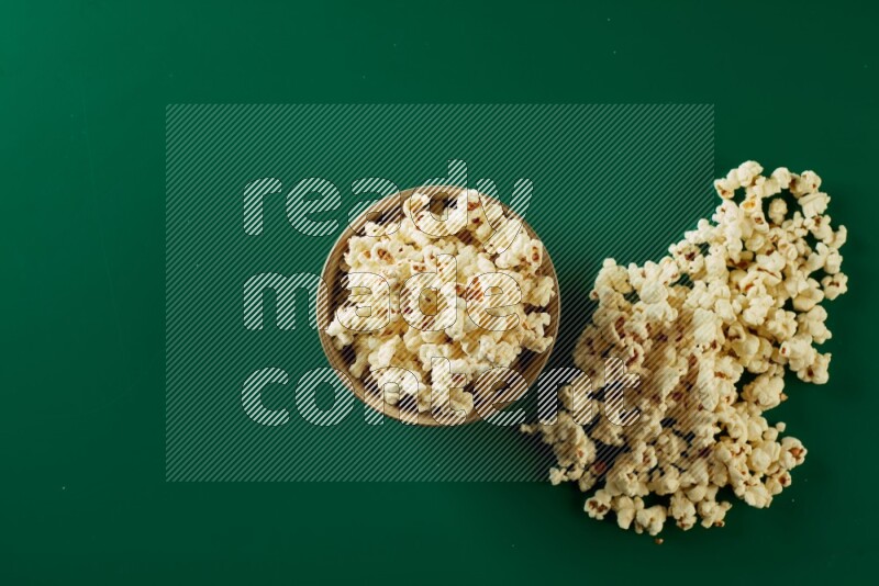 An off white ceramic bowl full of popcorn with popcorn beside it on a green background in a top view shot