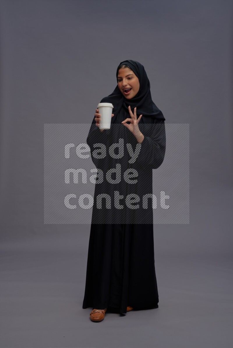 Saudi woman wearing Abaya standing holding paper cup on gray background