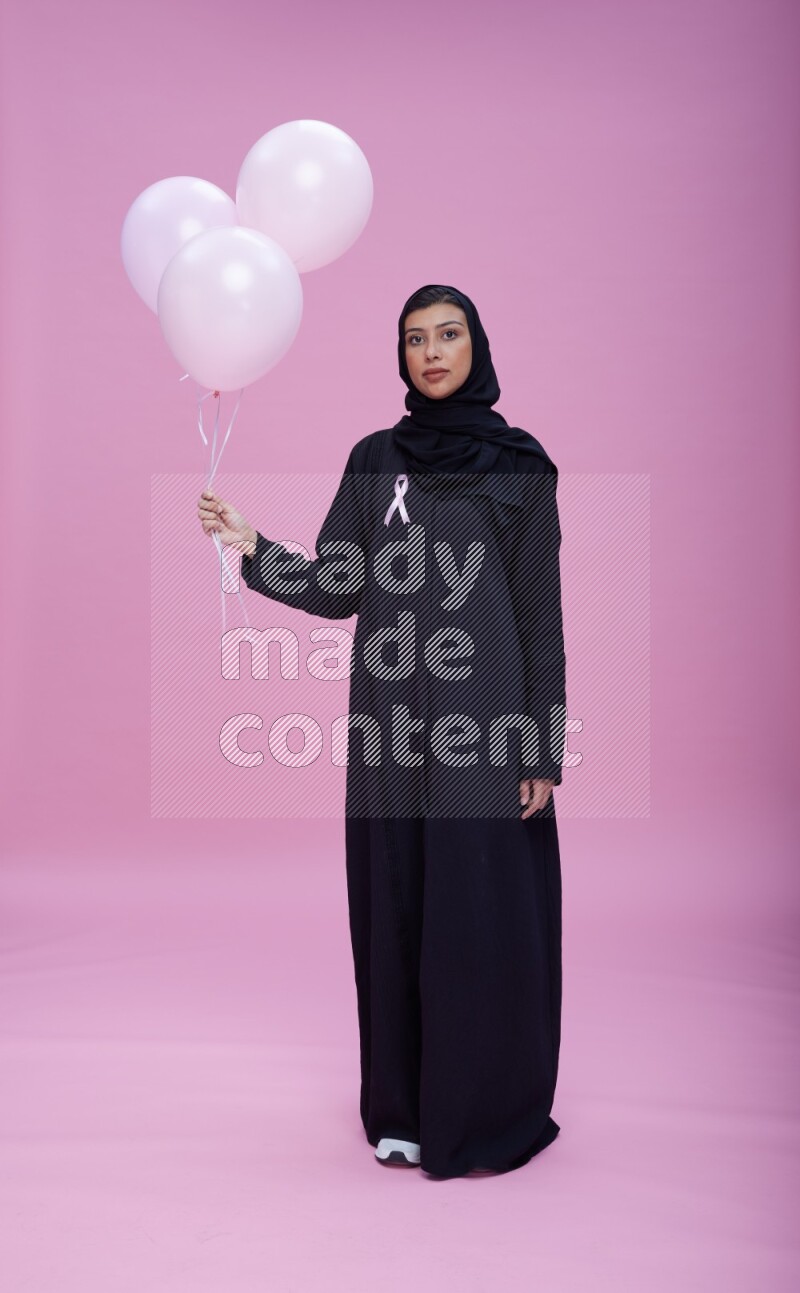 Saudi woman wearing pink ribbon on Abaya standing holding balloons on plain pink background