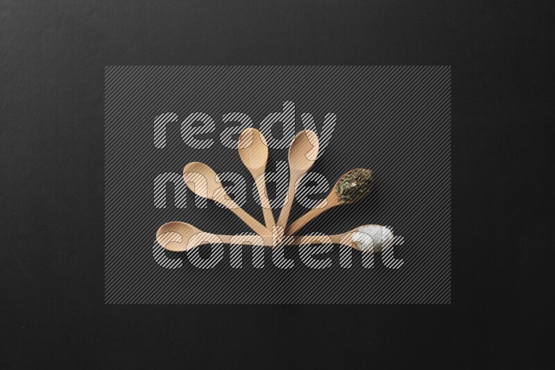 wooden spoons filled with white peppers, cloves, cardamom, salt, black peppers and basil on black flooring