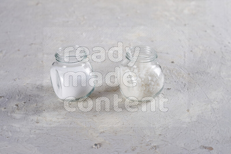 2 glass jars one is filled with table salt and the other with coarse sea salt on white background