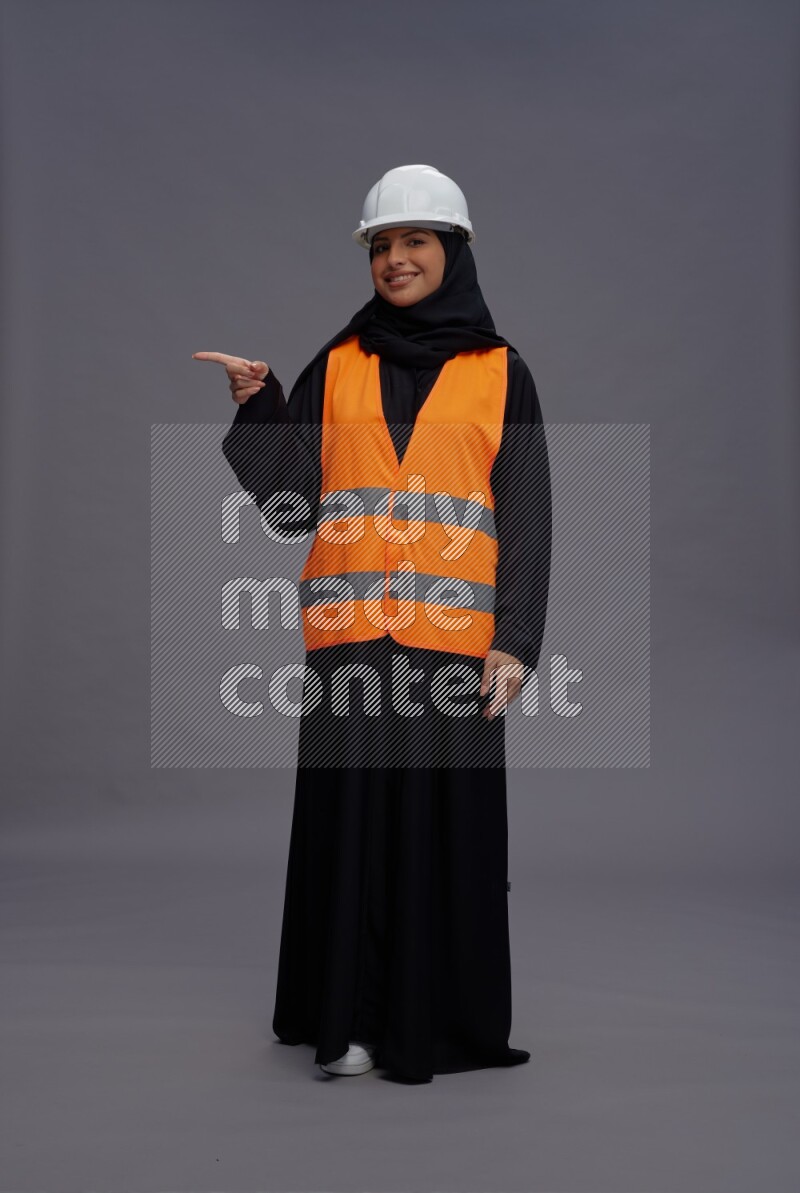 Saudi woman wearing Abaya with engineer vest standing interacting with the camera on gray background