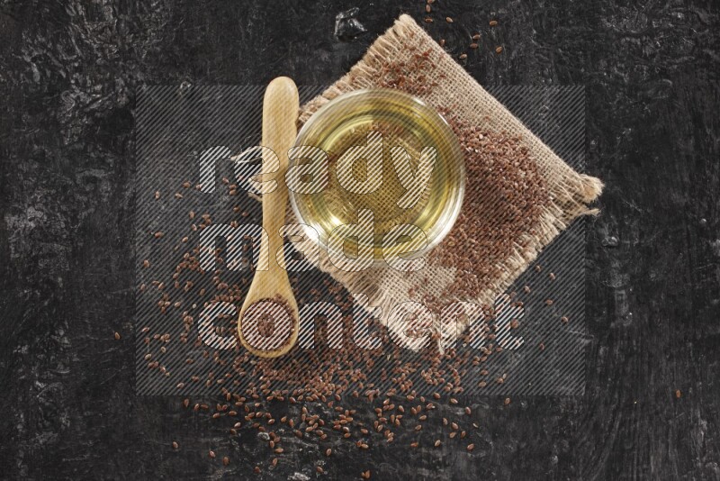 A glass bowl full of flaxseeds oil and wooden spoon full of flaxseeds with seeds spread on burlap fabric on a textured black flooring