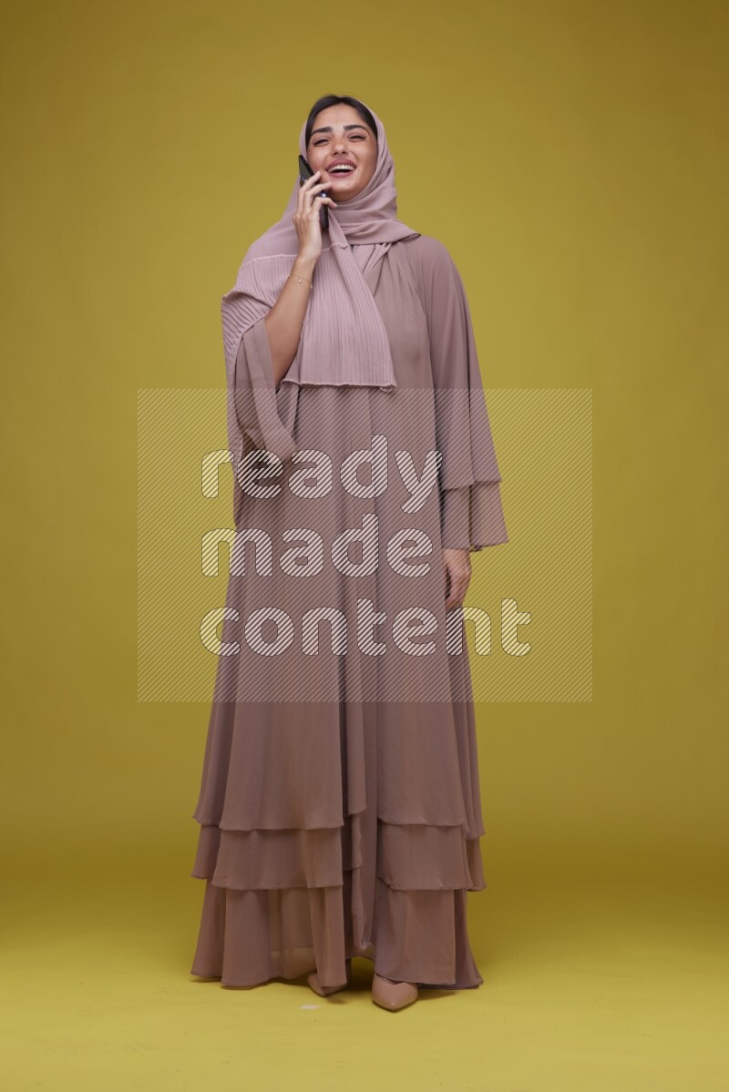 A woman having a call on a Yellow Background wearing Brown Abaya with Hijab