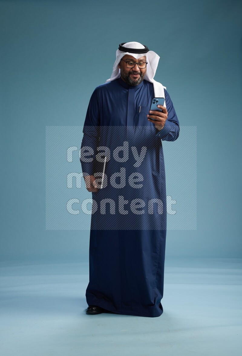 Saudi Man with shimag Standing texting on phone on blue background