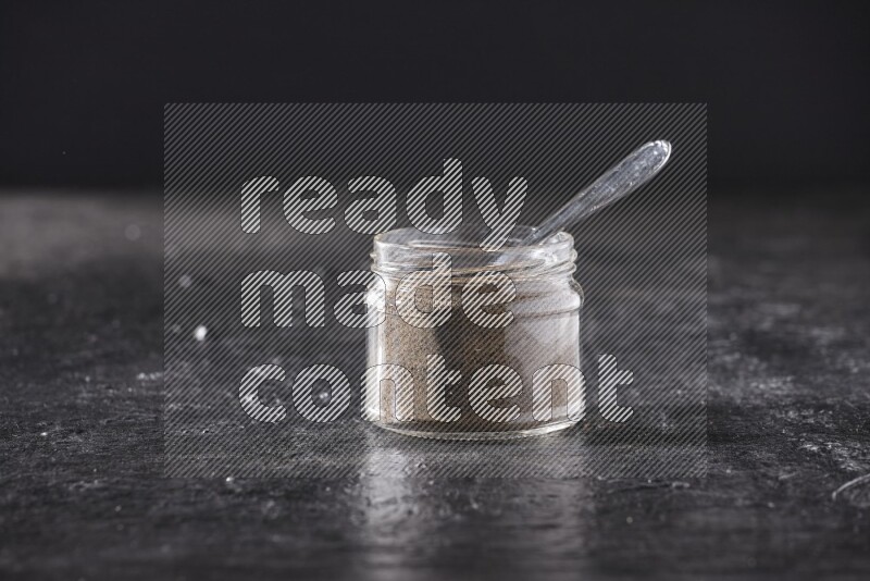 A glass jar full of black pepper powder and a metal spoon on a textured black flooring