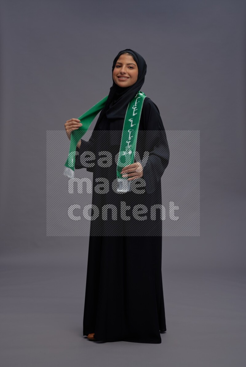 Saudi woman wearing Abaya standing holding Saudi flag on gray background