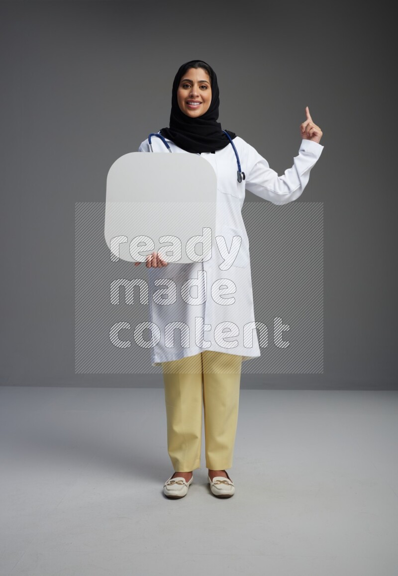 Saudi woman wearing lab coat with stethoscope standing holding social media sign on Gray background