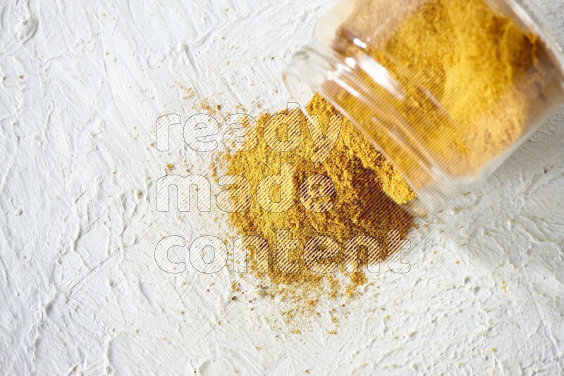 A flipped glass spice jar full of turmeric powder and powder fell out it on textured white flooring