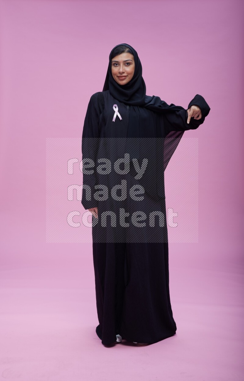 Saudi woman wearing pink ribbon on Abaya standing interacting with the camera on pink background
