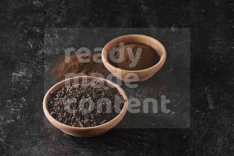 2 wooden bowls full of cloves powder and whole cloves on a textured black flooring