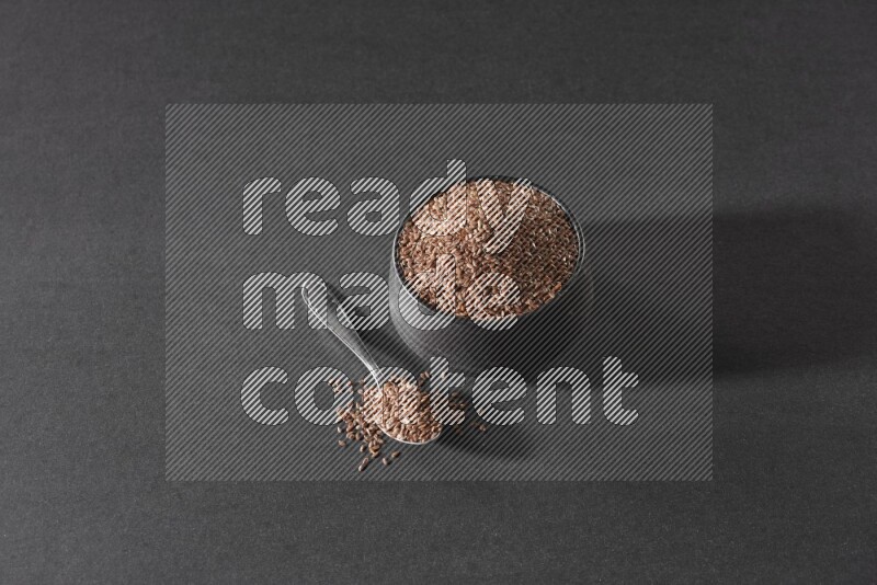 وعاء فخاري أسود ممتلئ بحبوب بذر الكتان مع ملعقة معدنية ممتلئة على أرضية سوداء