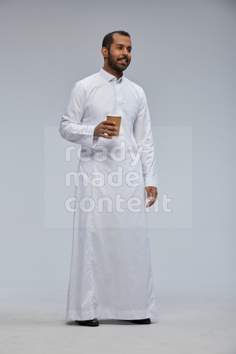 Saudi man wearing thob standing holding paper cup on gray background