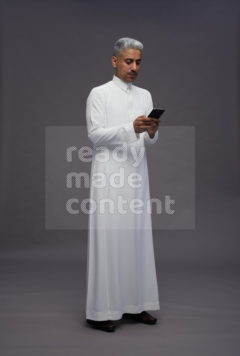 Saudi man wearing thob standing texting on phone on gray background