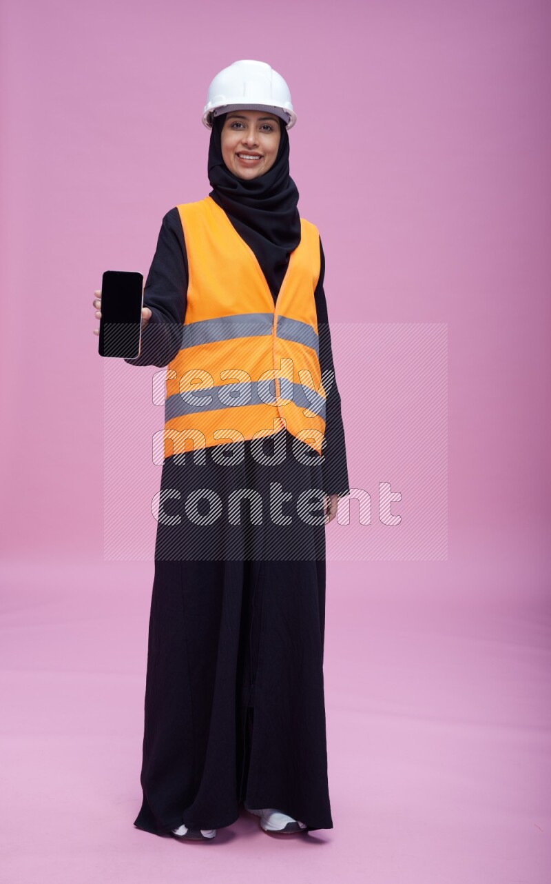 Saudi woman wearing Abaya with engineer vest and helmet standing showing phone to camera on pink background