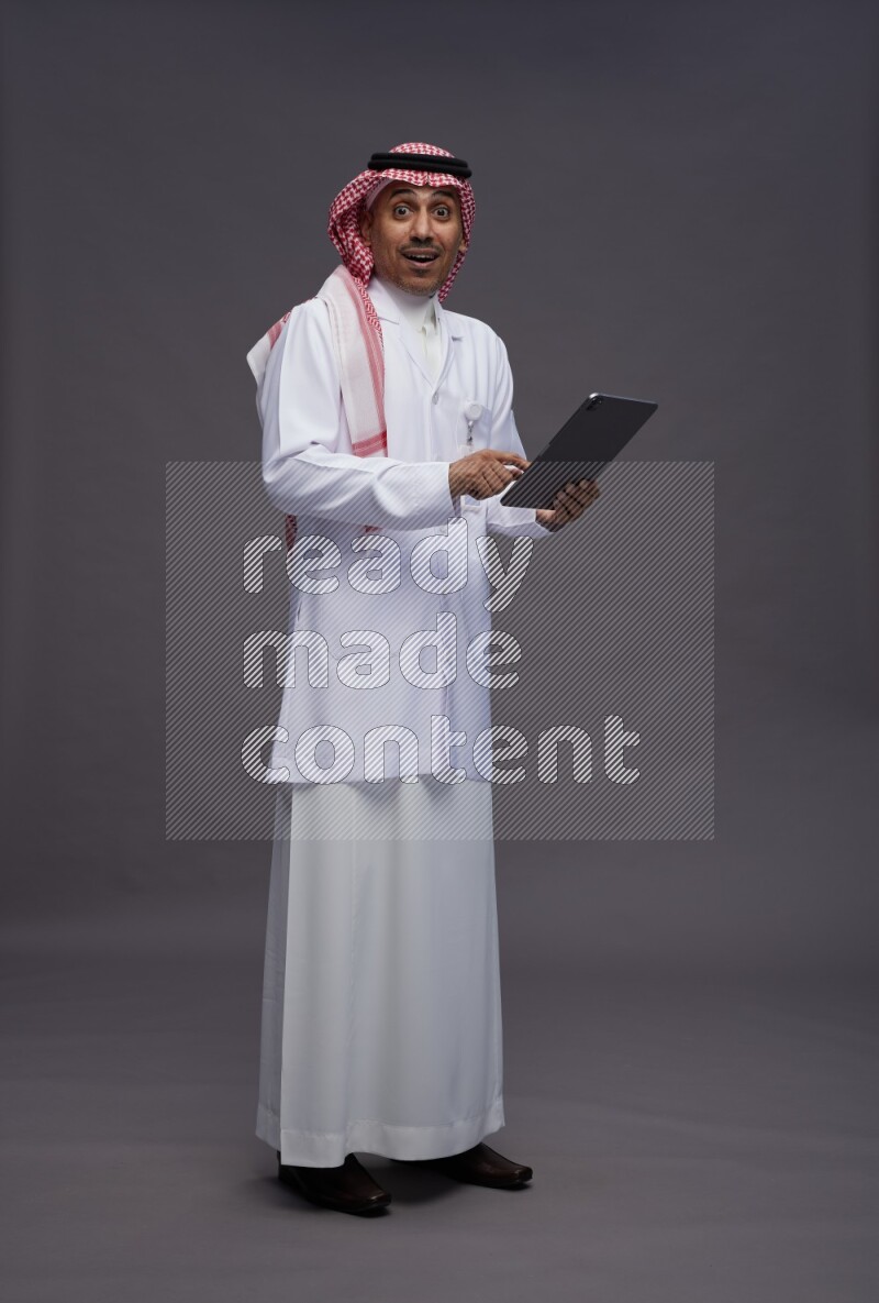 Saudi man wearing thob with lab coat and shomag with pocket employee badge standing working on tablet on gray background