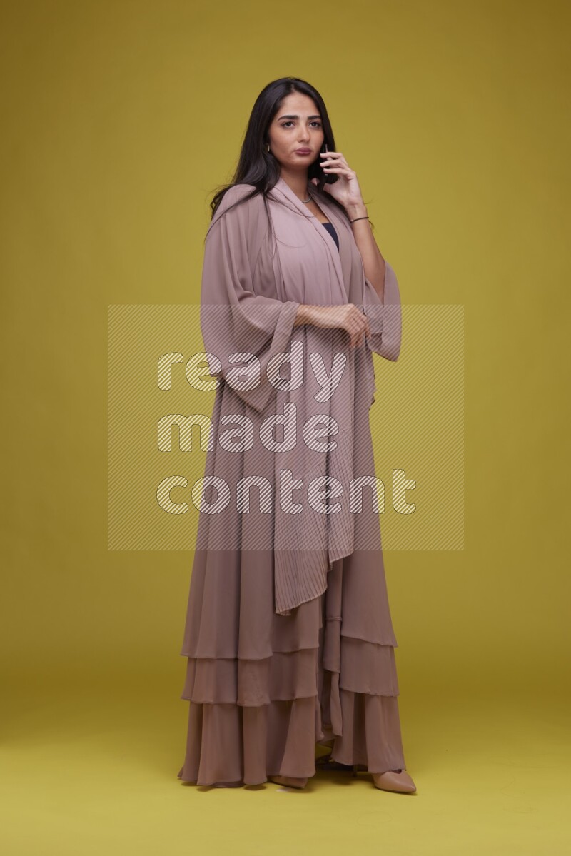 A woman Calling on a Yellow Background wearing Brown Abaya