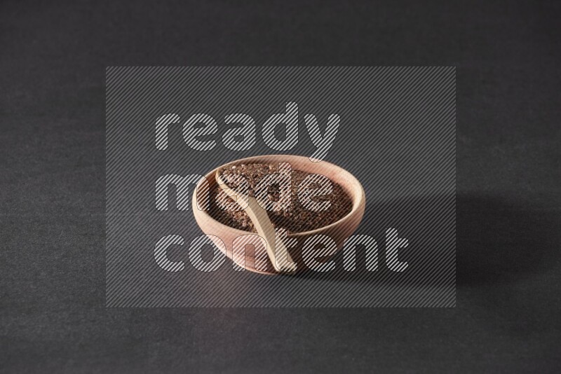 A wooden bowl full of flaxseeds with wooden spoon full of the seeds on it on a black flooring