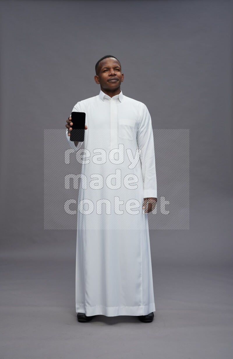 Saudi man wearing thob standing showing phone to camera on gray background