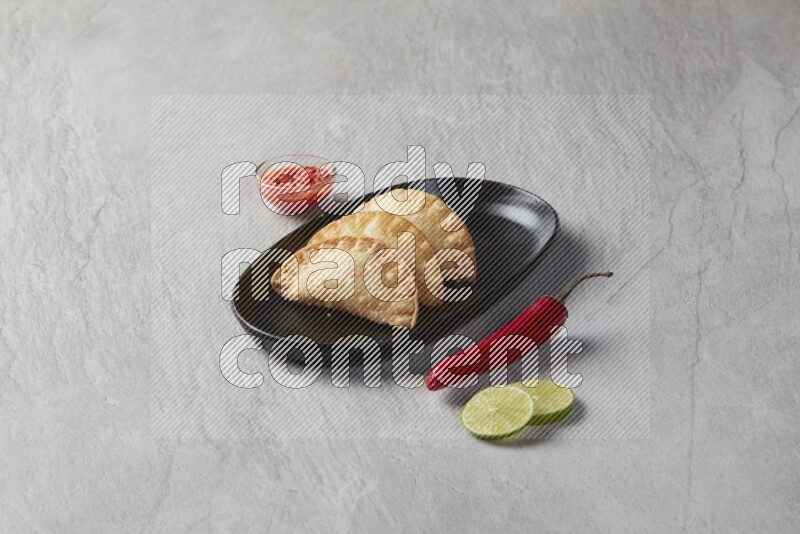 Three fried sambosas in an oval shaped black plate and a red sauce in a black round ramekin on a gray background