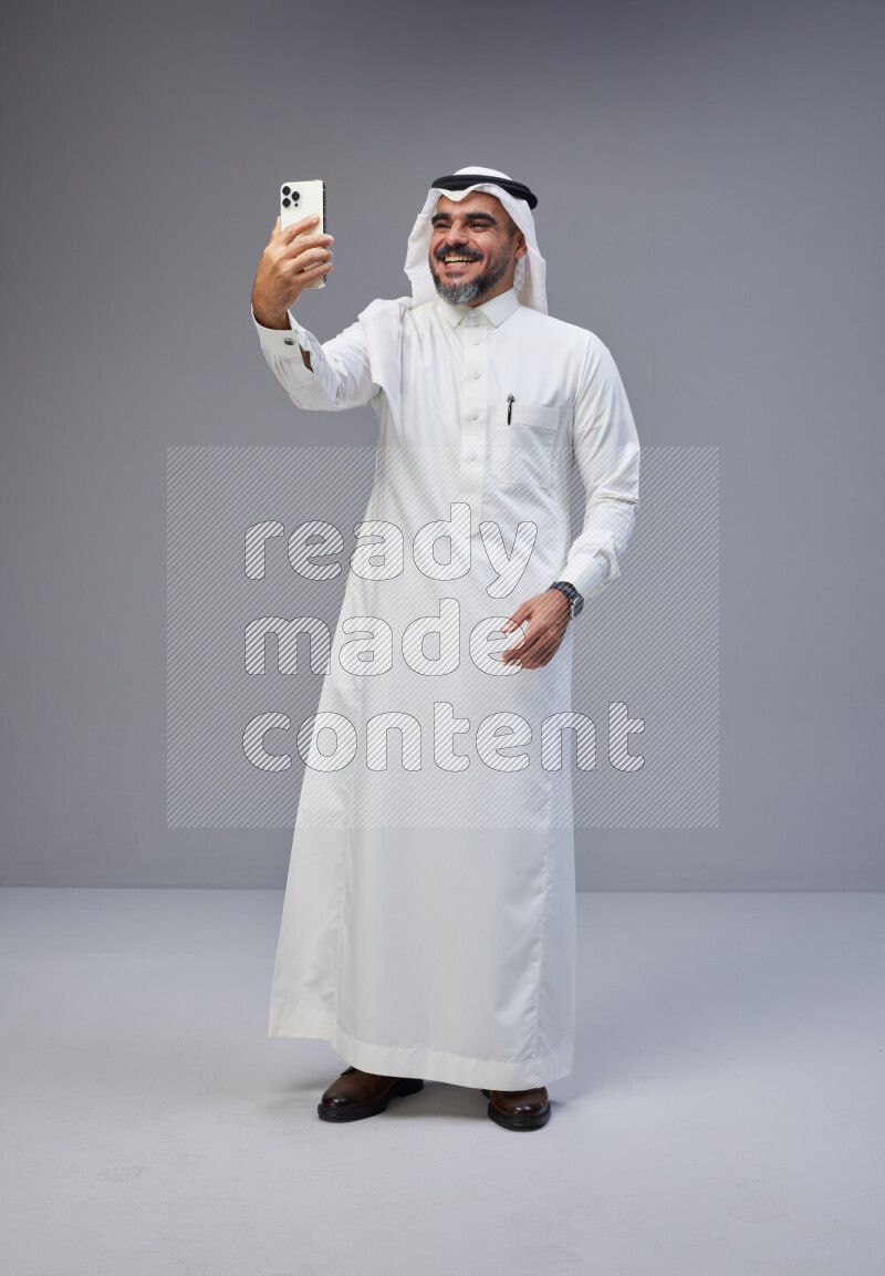 Saudi man Wearing Thob and white Shomag standing taking selfie on Gray background