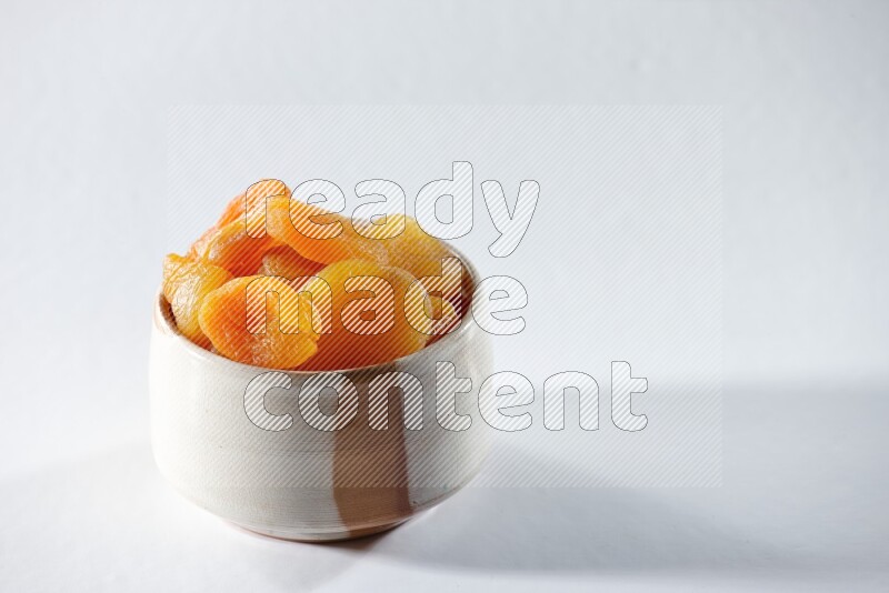 A beige ceramic bowl full of dried apricots on a white background in different angles