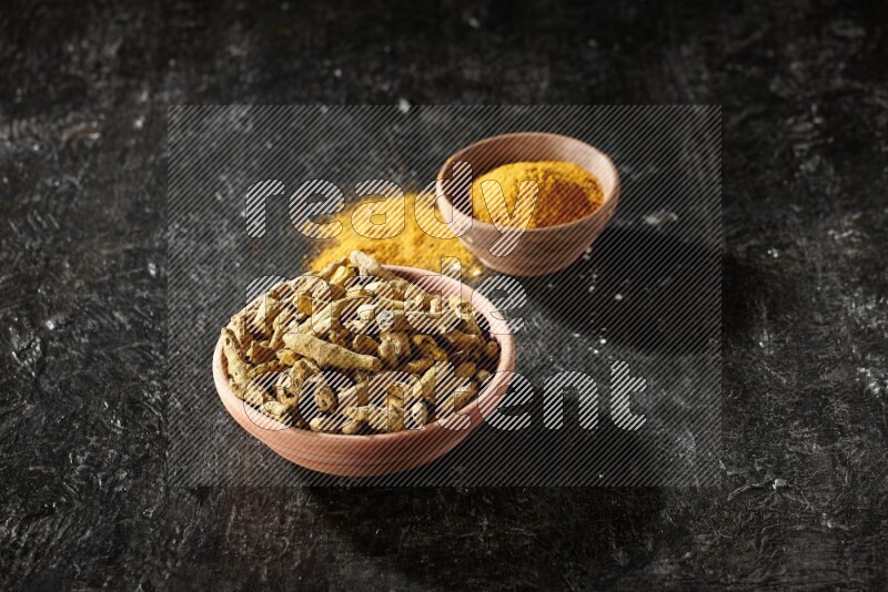 2 wooden bowls, one full of turmeric powder and the other full of dried turmeric whole fingers on a textured black flooring