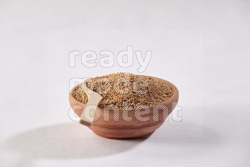 A wooden bowl full of mustard seeds with a wooden spoon on the seed on a white flooring