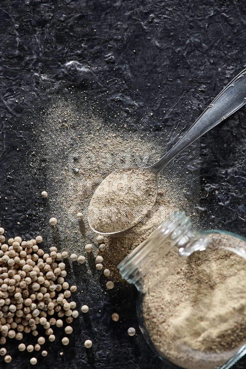 A flipped herbal glass jar and metal spoon full of white pepper powder with spilled powder and pepper beads on textured black flooring