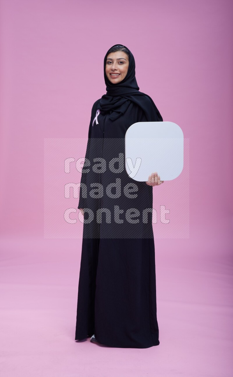 Saudi woman wearing pink ribbon on Abaya standing holding social media signs on pink background