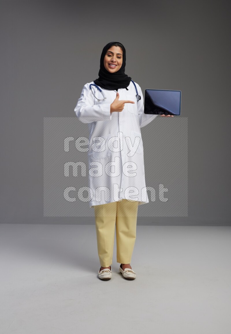 Saudi woman wearing lab coat with stethoscope standing showing tablet to camera with sign in the back on Gray background