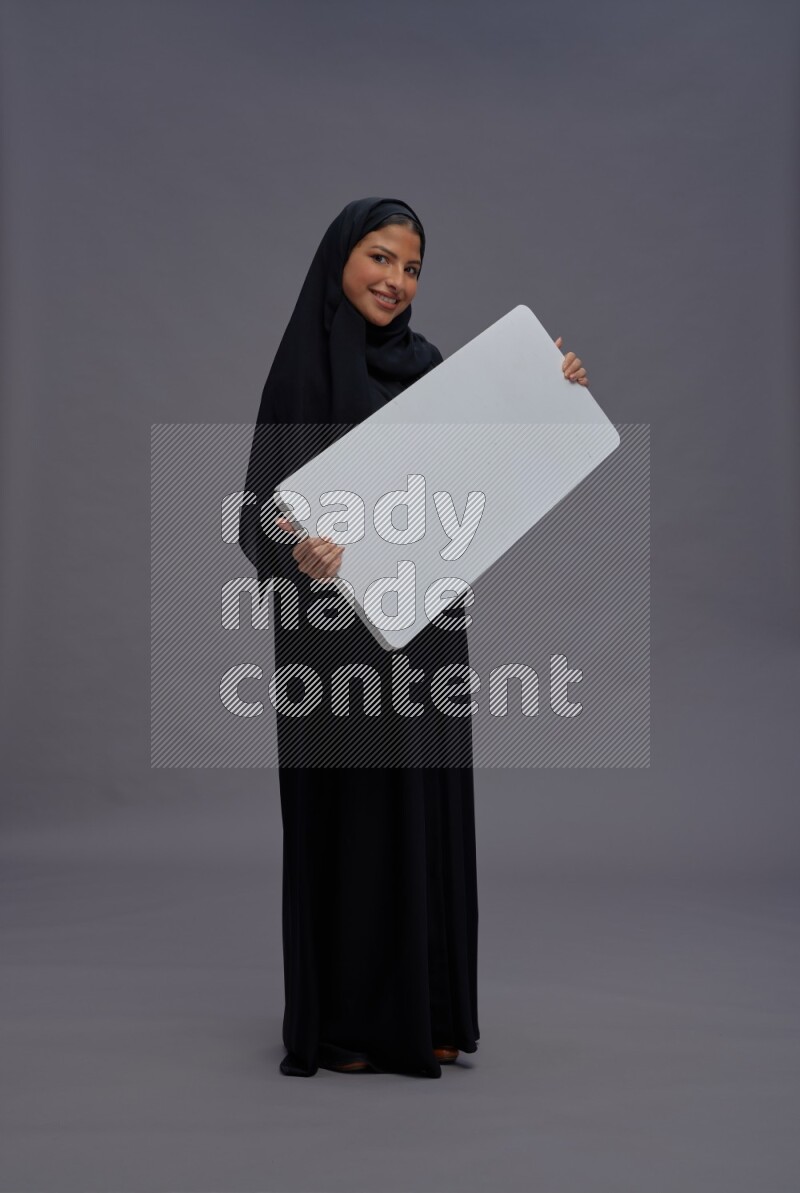 Saudi woman wearing Abaya standing holding white board on gray background