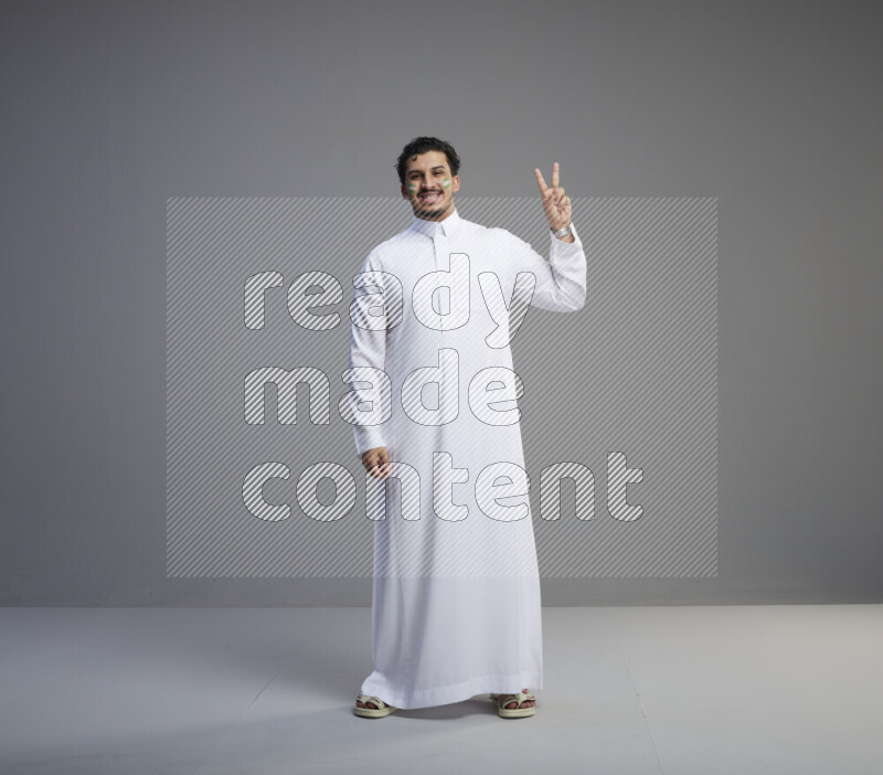 A Saudi man standing wearing thob with face painting interacting with the camera on gray background