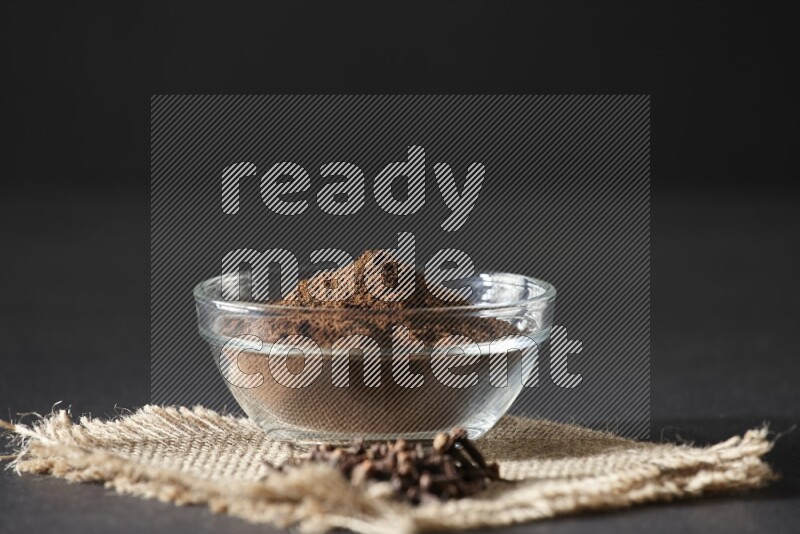 A glass bowl full of cloves powder with cloves grains on a burlap piece on a black flooring