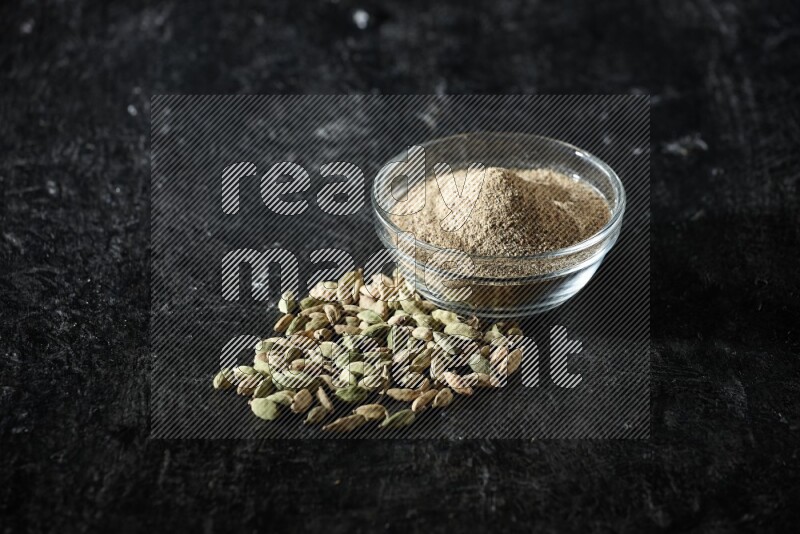 A glass bowl full of cardamom powder and cardamom seeds beside it on textured black flooring