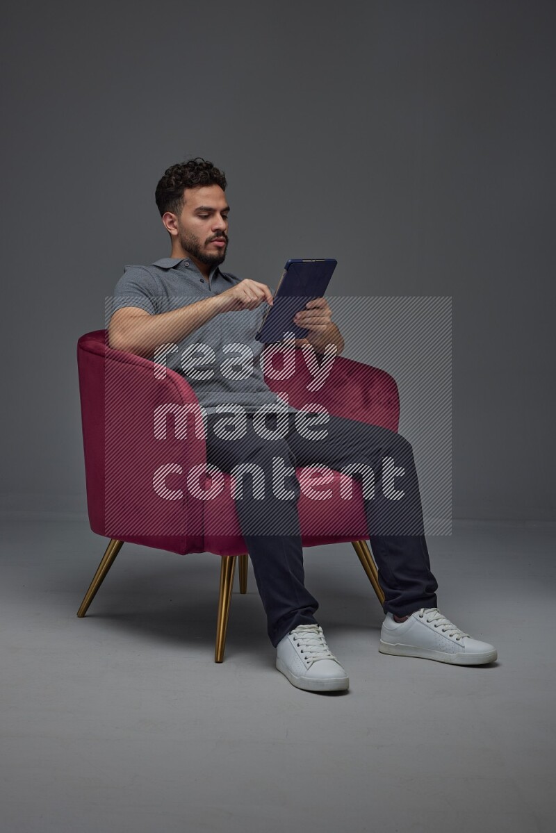 A man wearing casual and using his tablet while sitting on a burgundy chair eye level on a gray background