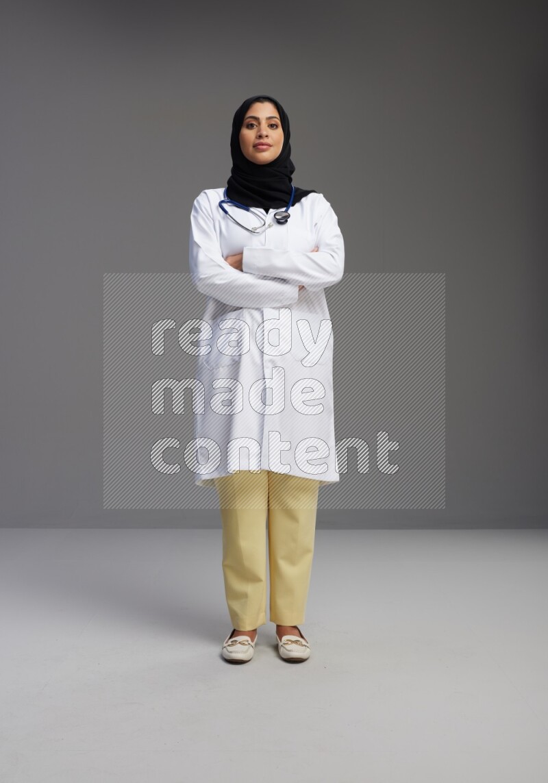 Saudi woman wearing lab coat with stethoscope standing with crossed arms on Gray background