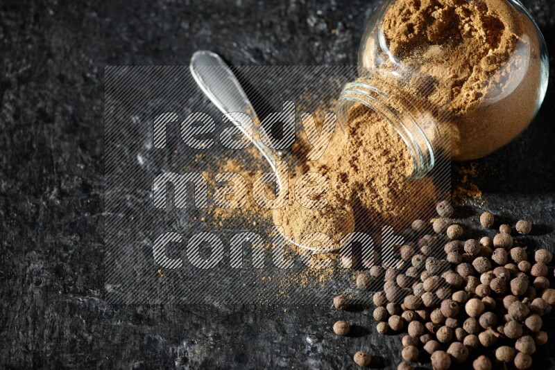 A flipped glass spice jar and metal spoon full of allspice powder and powder spilled out of it with whole balls on a textured black flooring