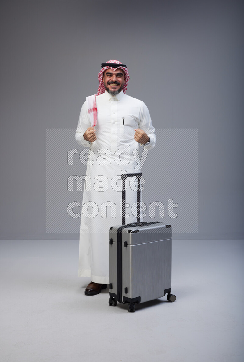 Saudi man wearing Thob and red Shomag standing holding Travel bag on Gray background
