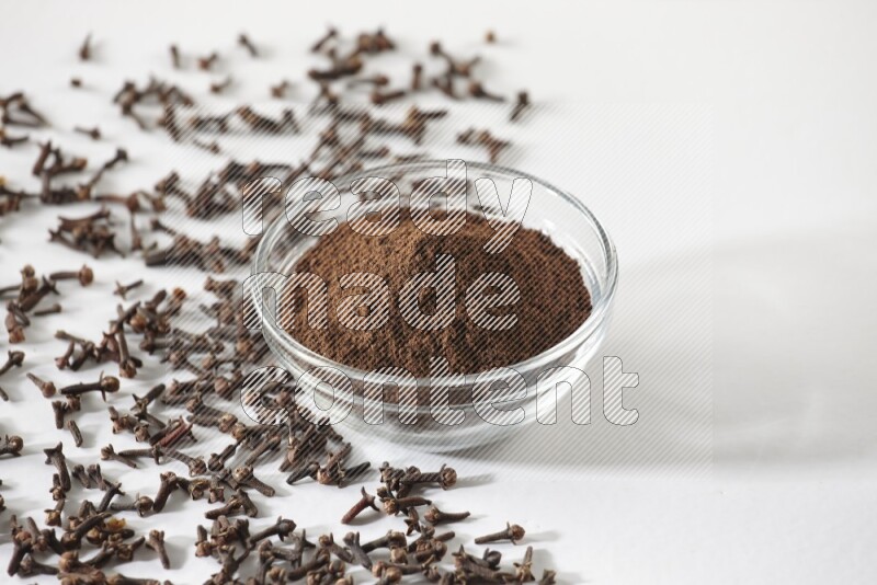 A glass bowl full of cloves powder and cloves grains spread on white flooring