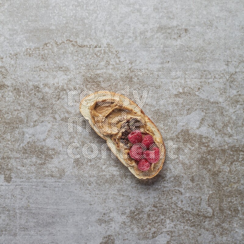 open faced peanut butter sandwich with raspberries and chocolate granula on a grey textured background