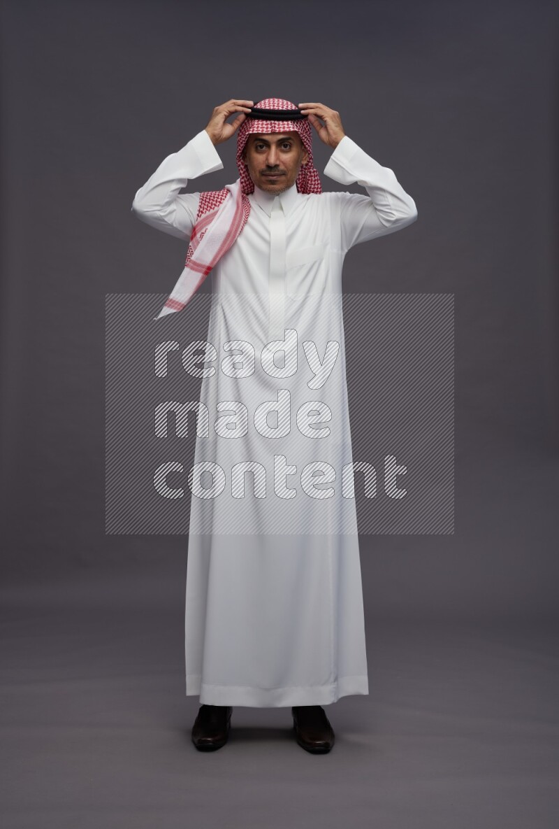 Saudi man wearing thob and shomag standing hands behind head on gray background