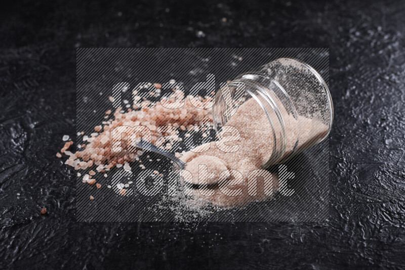 A glass jar full of fine himalayan salt with some himalayan crystals beside it on a black background
