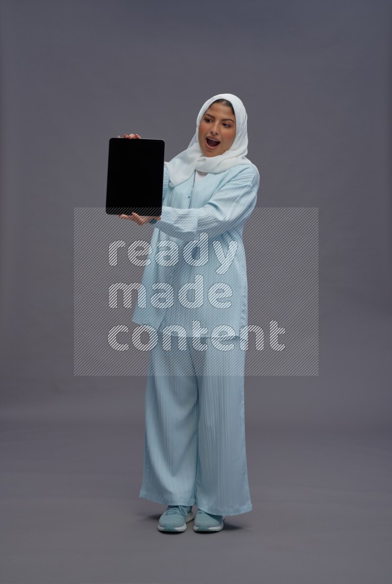 Saudi woman wearing hijab clothes standing showing tablet to camera on gray background